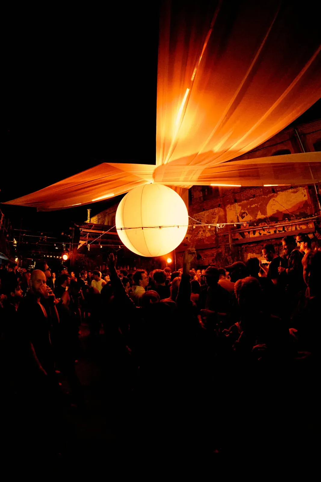 Sphère décorative lumineuse lors d’un événement de musique électronique Sphère décorative lumineuse suspendue au-dessus d’une piste bondée, mur de brique et drapés en arrière-plan.
