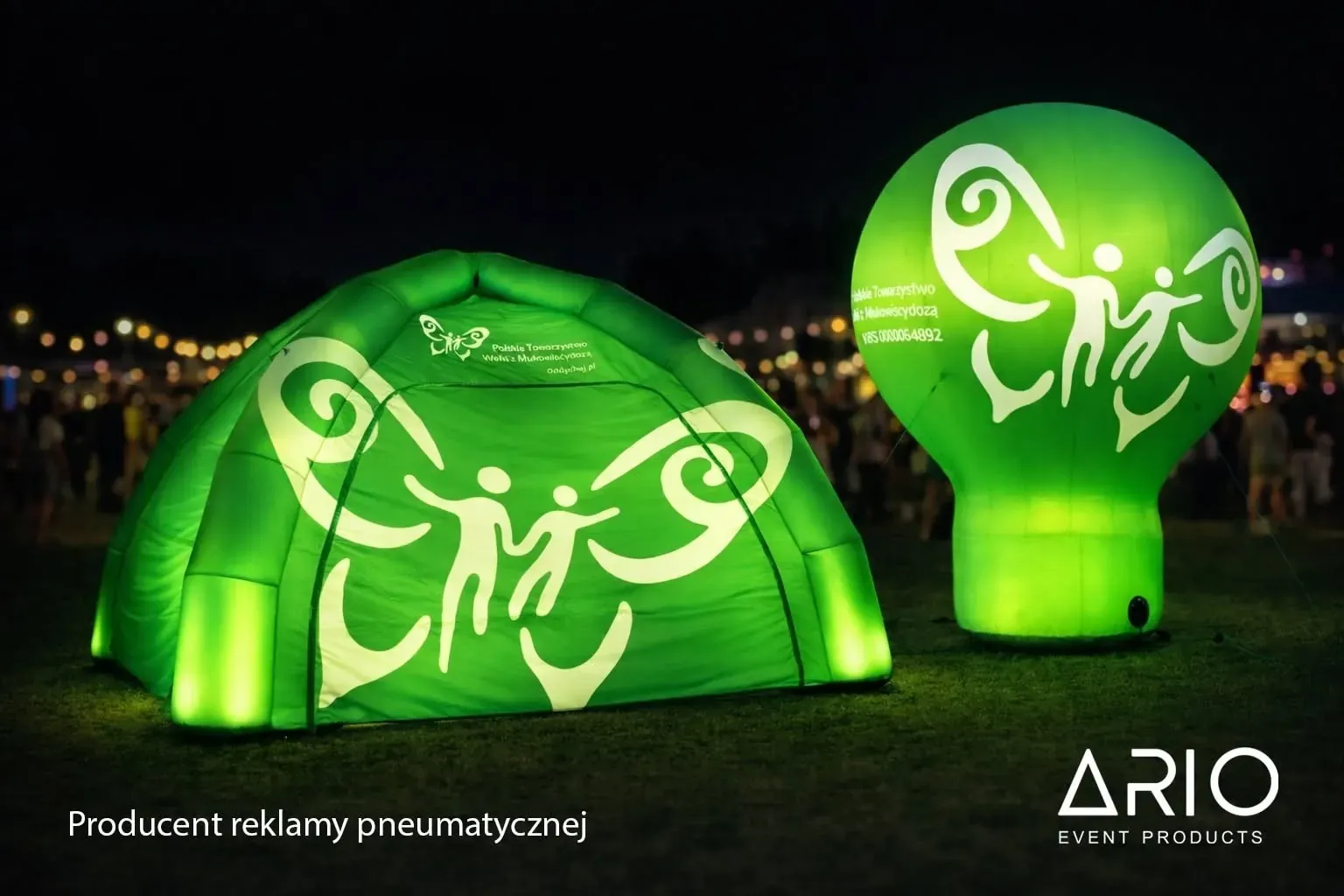 Namiot pneumatyczny i balon reklamowy rozświetlone na zielono po zmroku, w tle tłum na imprezie plenerowej.